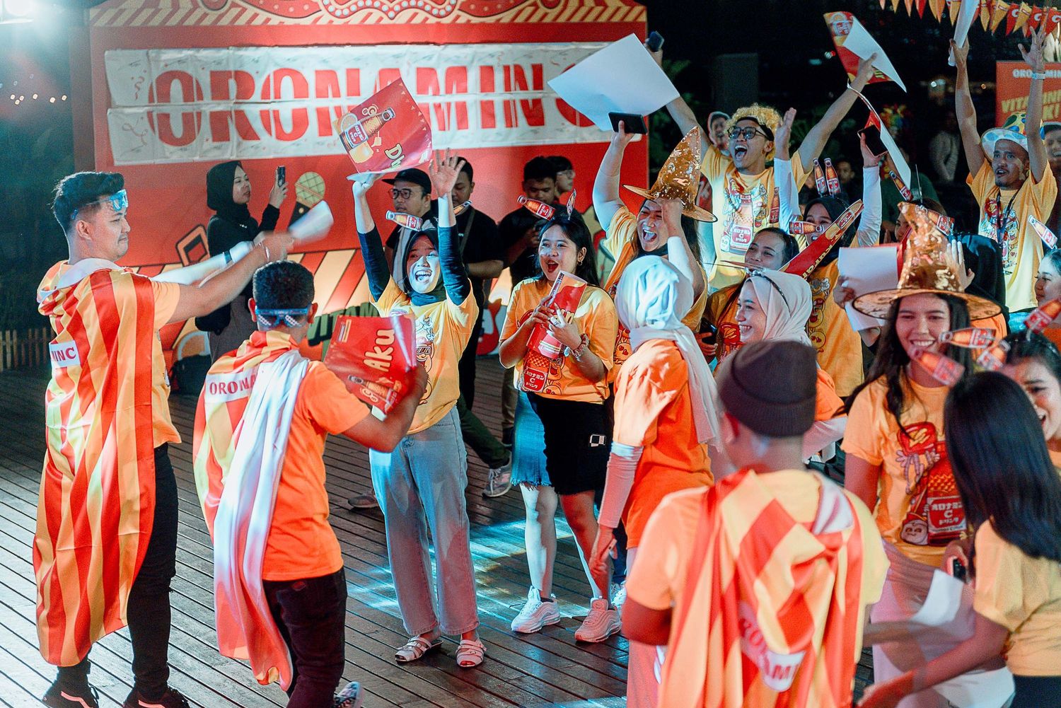 Energetic crowd celebrates at an event wearing orange and white attire while dancing and cheering together.