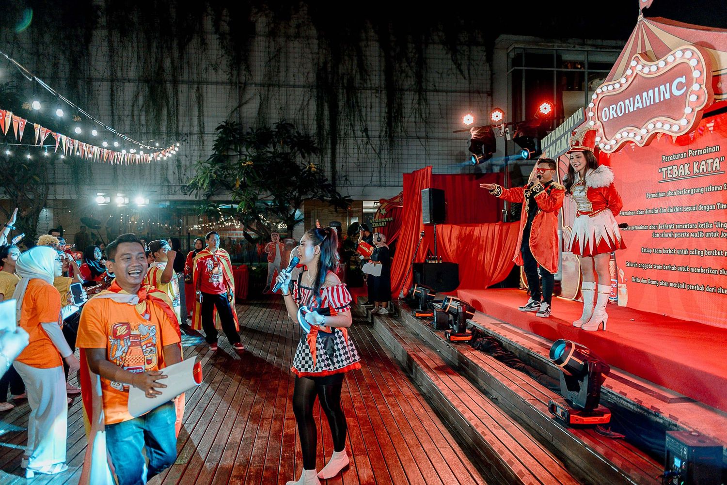 People gather at a nighttime event with red stage lighting and string lights creating a festive outdoor atmosphere.
