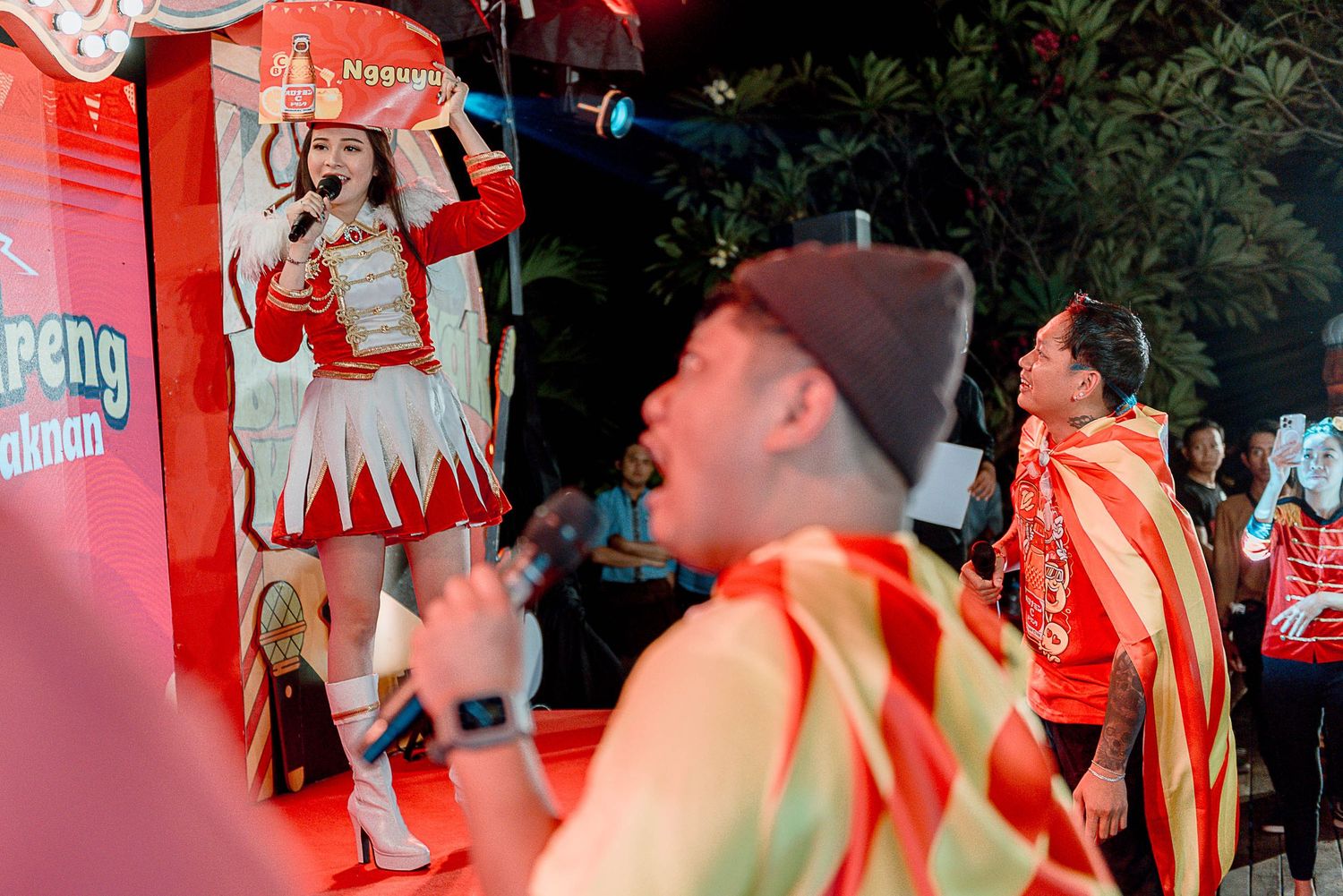 A figure in red and white medieval costume performs at an outdoor event with red decorative elements in the background.