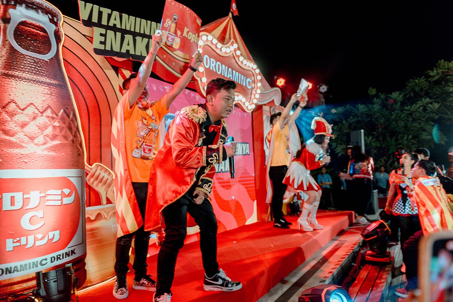 Performers on a red-lit stage with large Vitamin Energy drink promotional displays and decorative elements in the background.