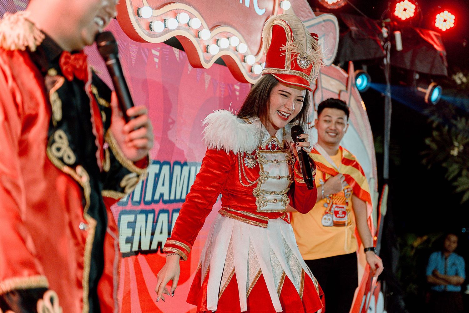 Performer in red and white marching band uniform with feathered hat dances during festive celebration with colorful lights.