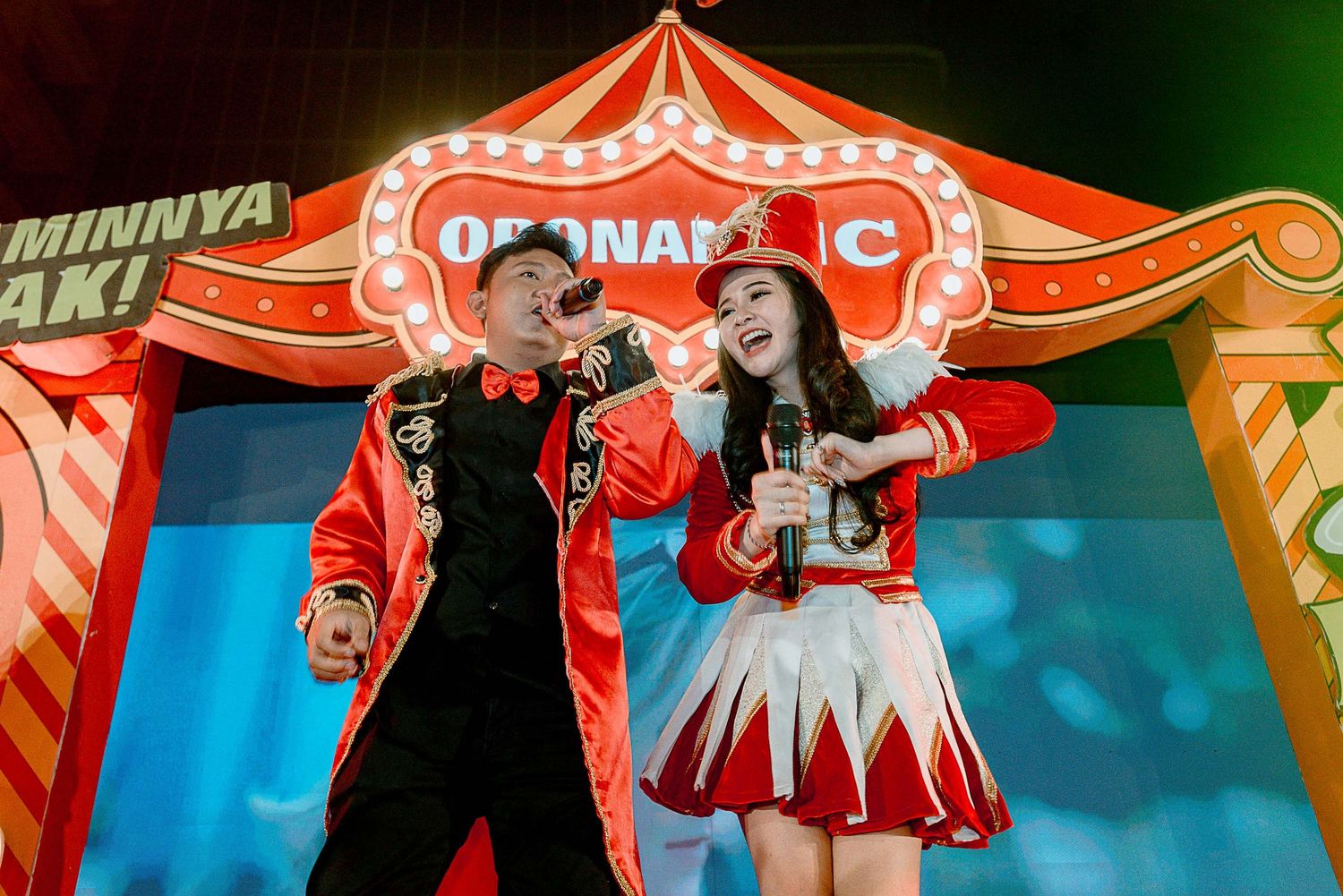 Performers in red and black costumes dance and interact on a colorful stage with decorative banners and lighting.
