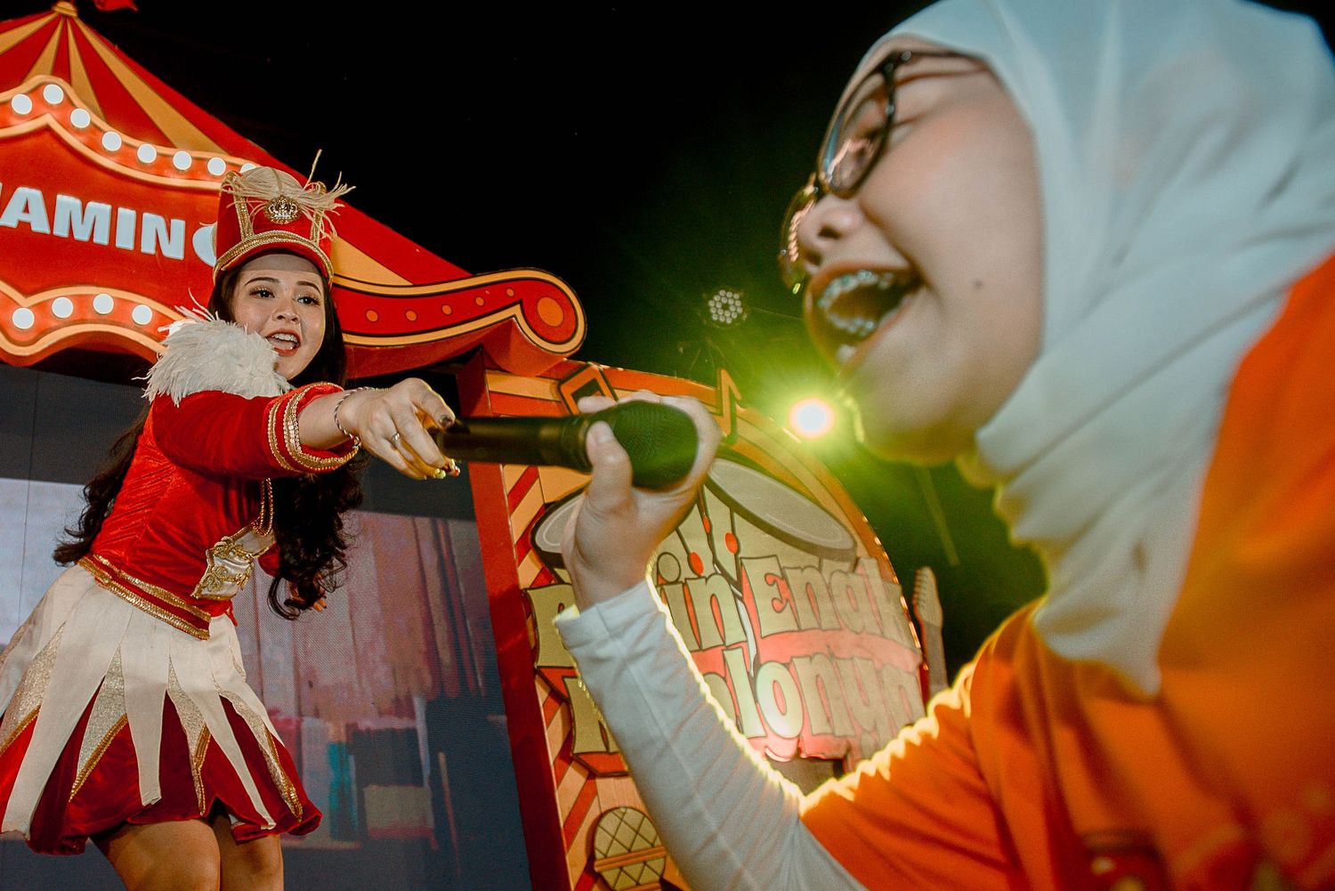 Two people share a joyful moment at a carnival with red and orange lighting and decorative signage in the background.