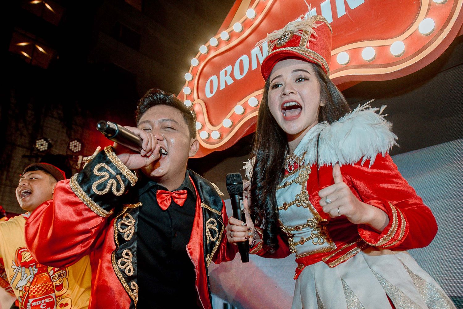 Entertainers in theatrical circus-style costumes performing with animated expressions under red lighting.