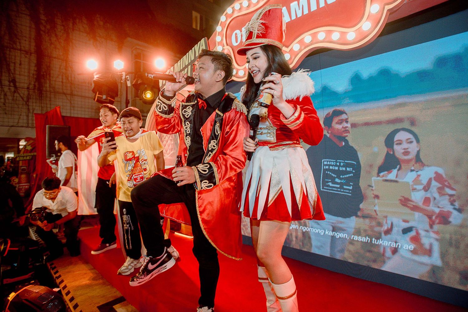 People in red and white outfits perform on stage at an entertainment venue with colorful lighting and video projection.
