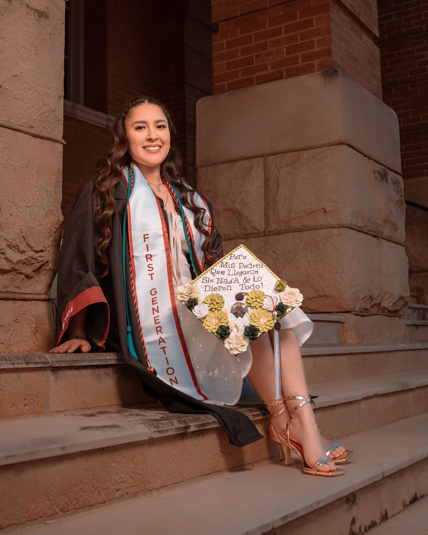 Graduate in a sash poses on concrete steps with yellow and white flowers against a brick building backdrop.