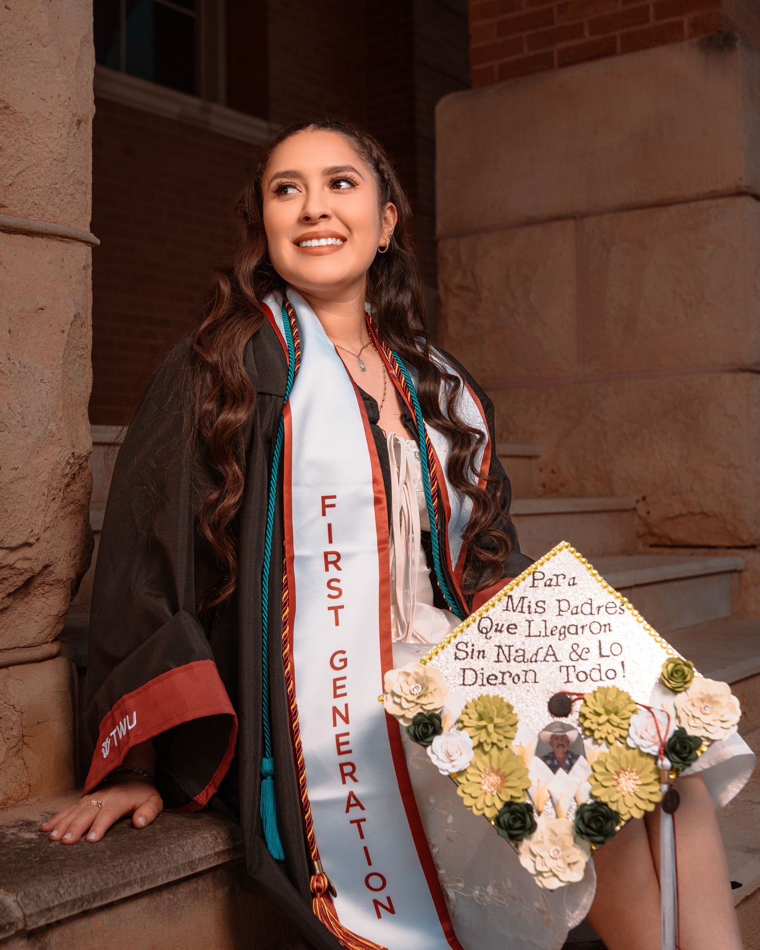 A graduate wearing a white sash and holding a decorated graduation cap poses in front of a brick building.