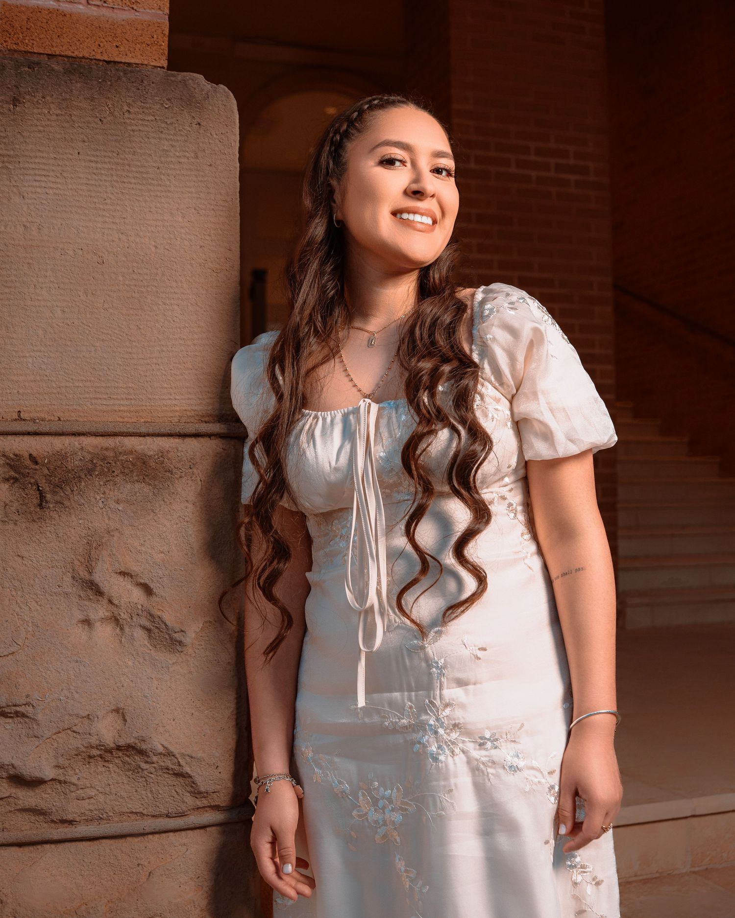 A woman in a vintage-style white dress poses against a rustic brick wall with warm lighting and long wavy hair.