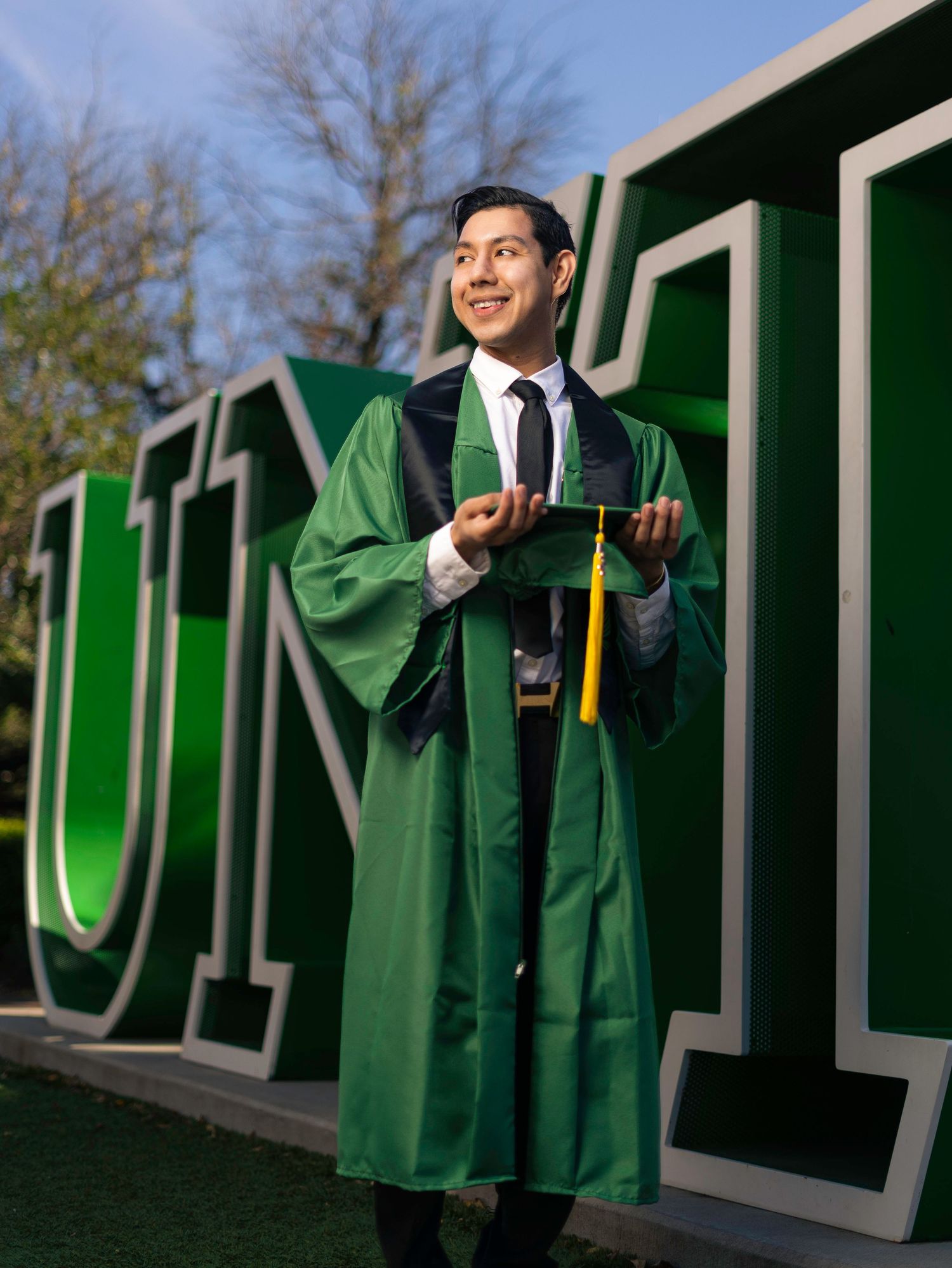 Graduate in green commencement gown poses with large outdoor university letters in a series of celebratory photos.