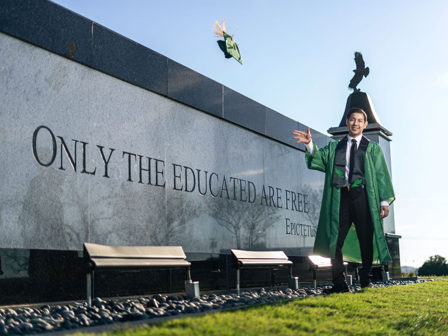 Graduates in green caps and gowns stand against a long concrete wall during an outdoor commencement ceremony.