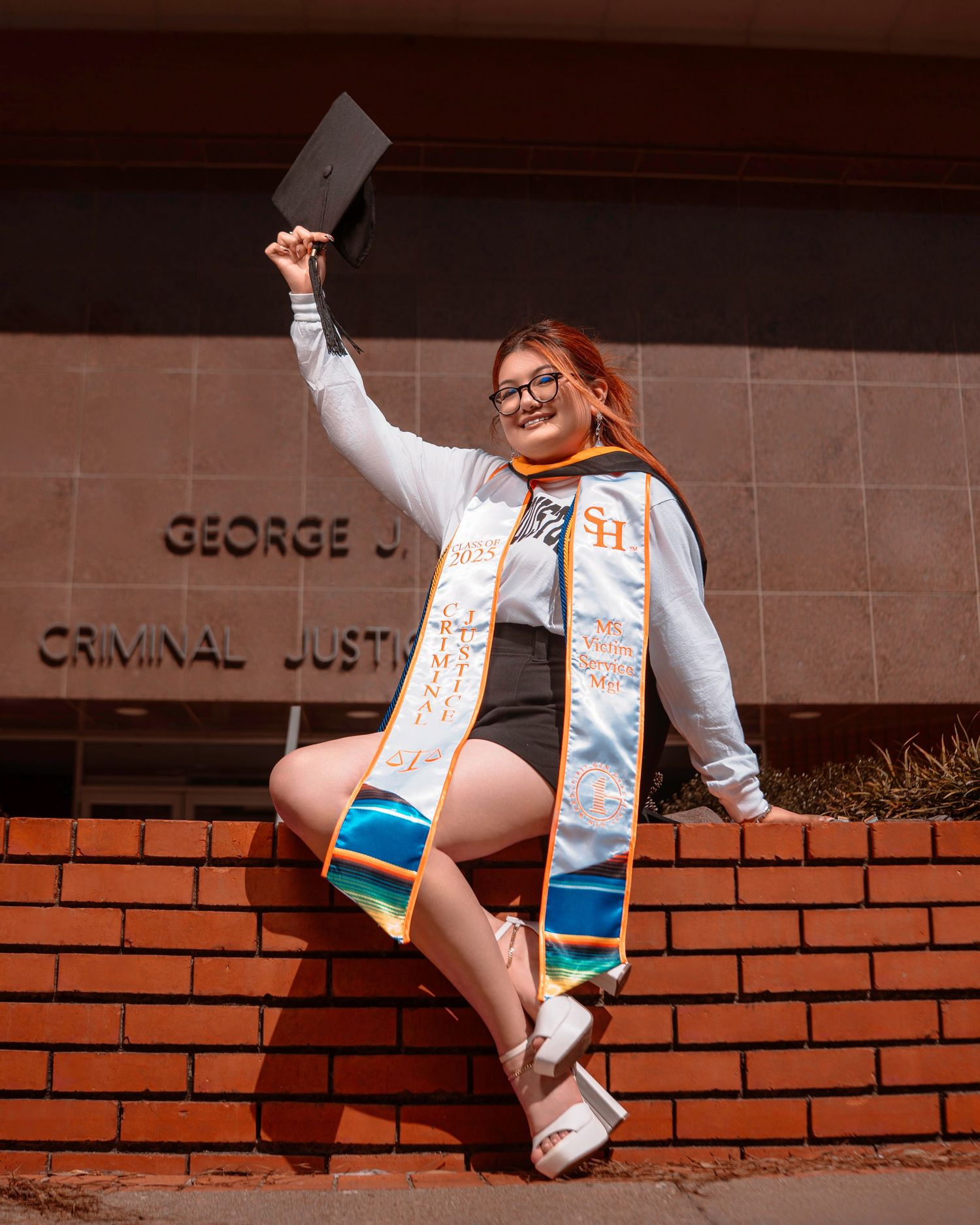 Graduate celebrates on brick wall outside Criminal Justice building wearing decorated graduation stole and holding diploma.