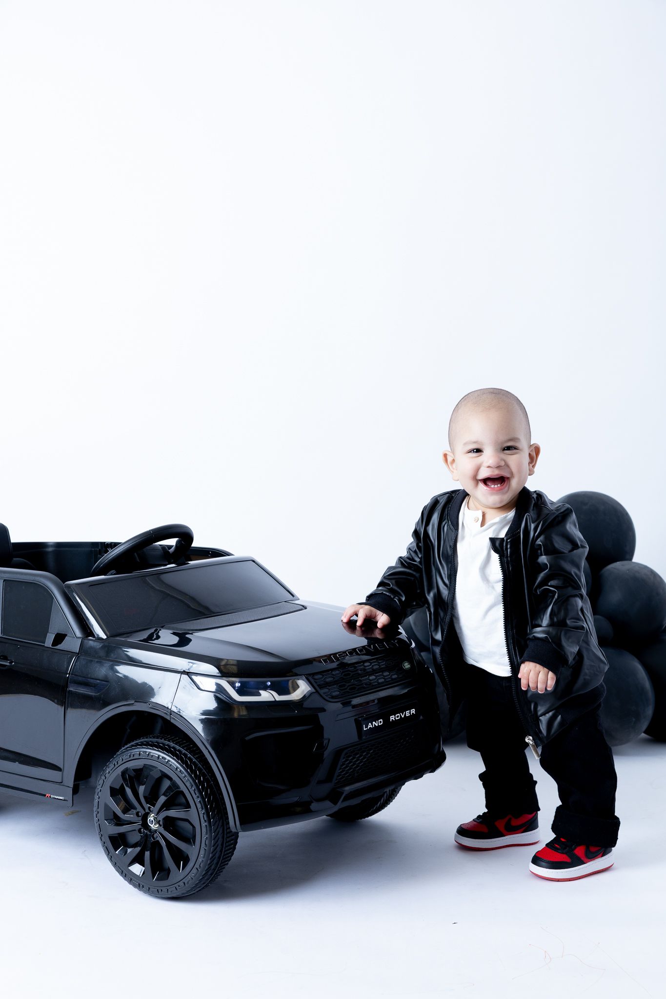 Baby in black leather jacket and sneakers poses next to mini electric car with black balloons.
