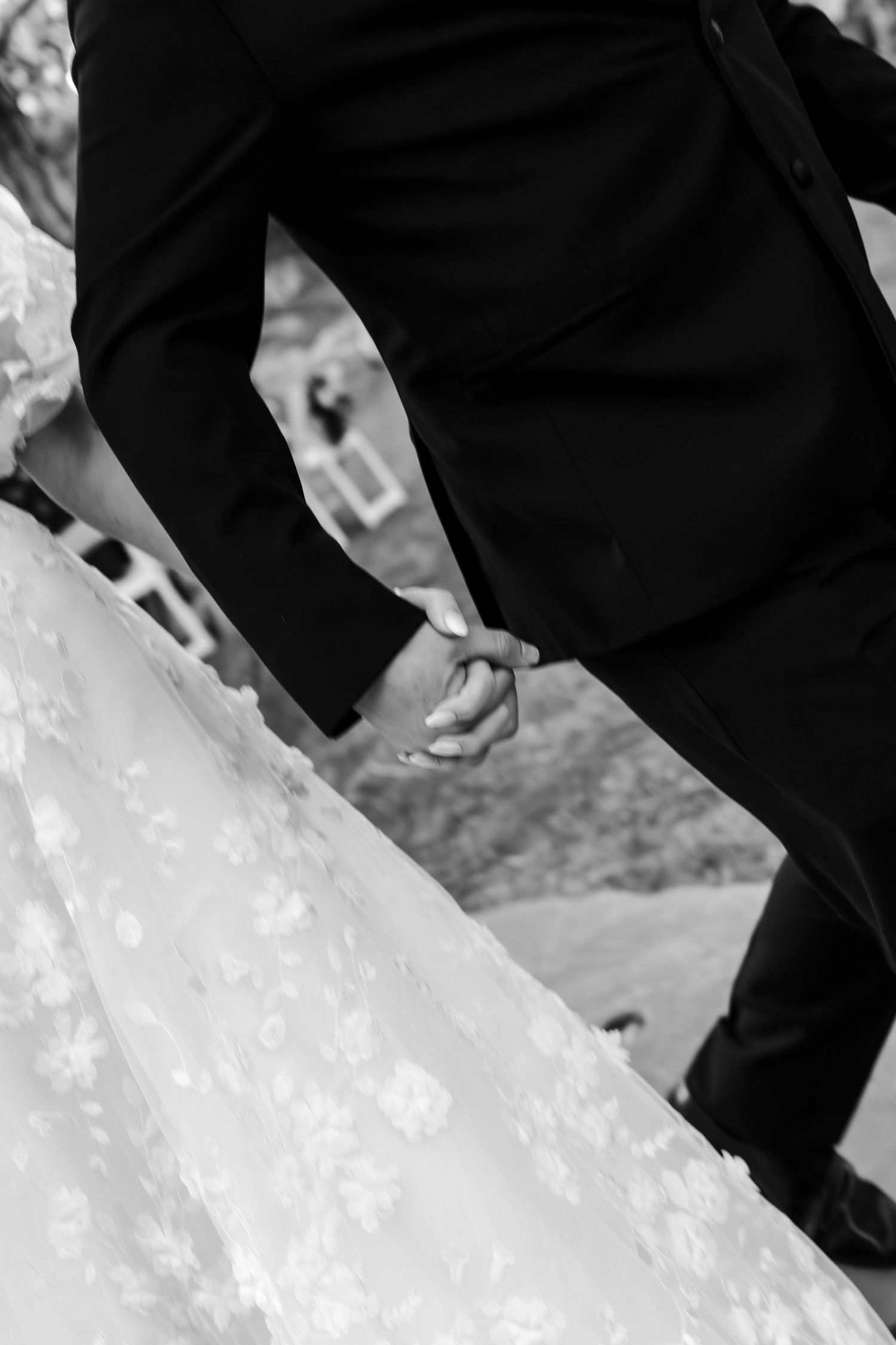 Black and white close-up of a bride and groom holding hands, showcasing her lace wedding gown during the ceremony.