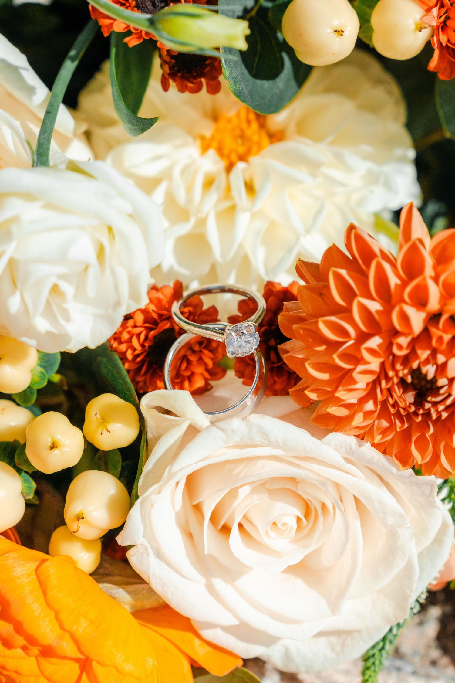 Diamond engagement ring nestled in bridal bouquet of white roses and orange dahlias.
