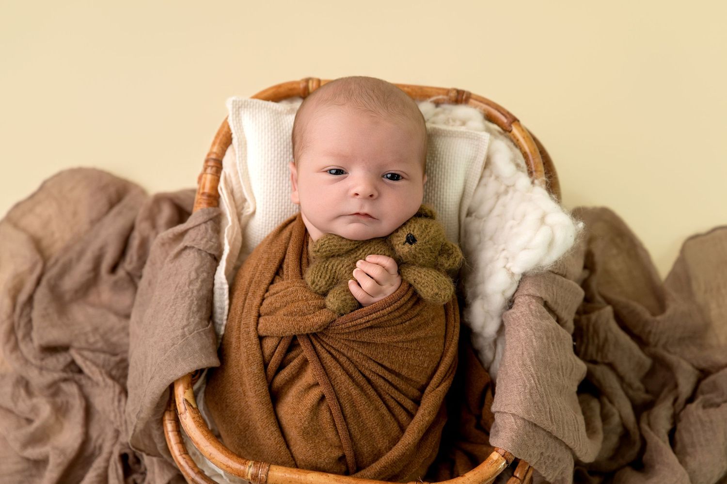 A baby wrapped in brown swaddle blanket lies on beige bedding with matching earth-toned decor.