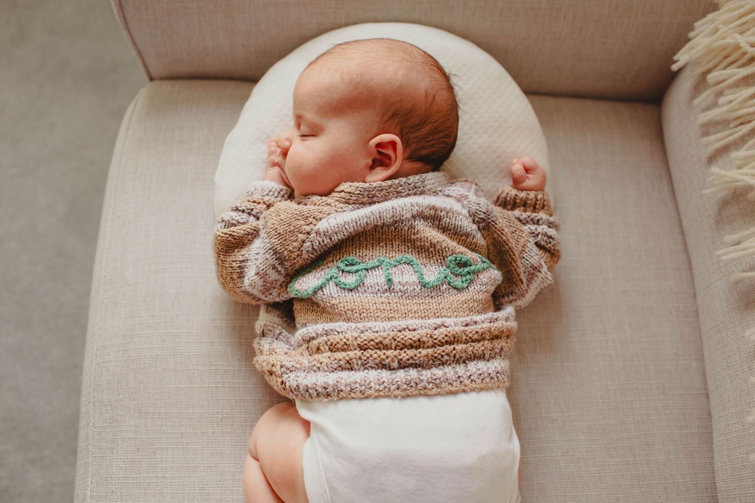 Newborn baby sleeping peacefully on beige linen couch wearing soft knit sweater.