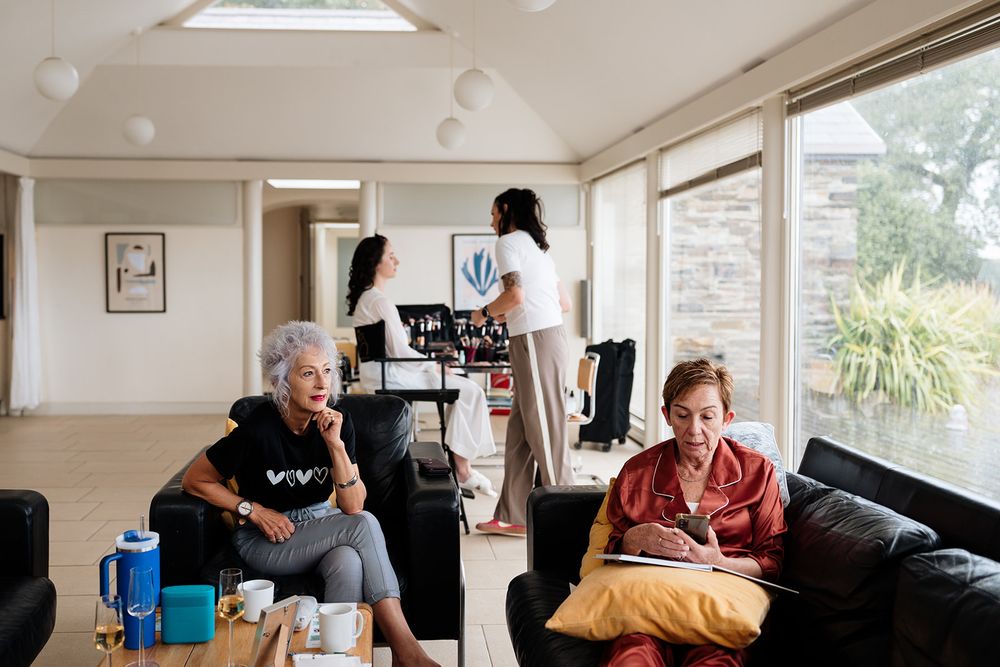People relax in a bright living room with large windows while someone works in the background.