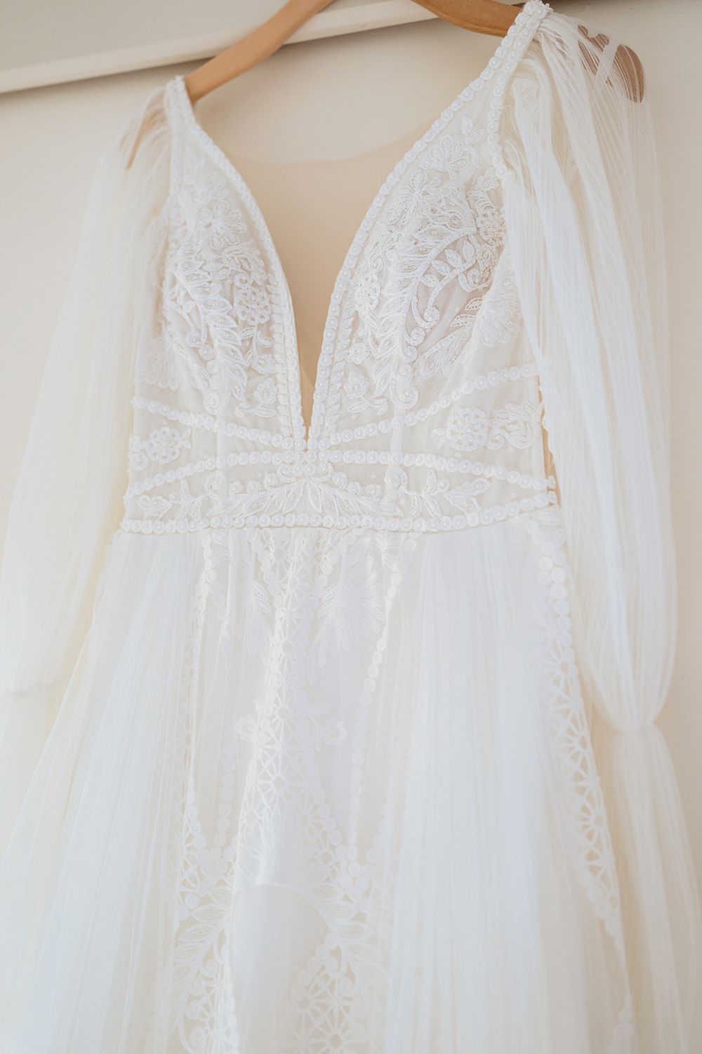 White lace wedding dress with plunging neckline and delicate floral details hanging on wooden hangers against beige wall.
