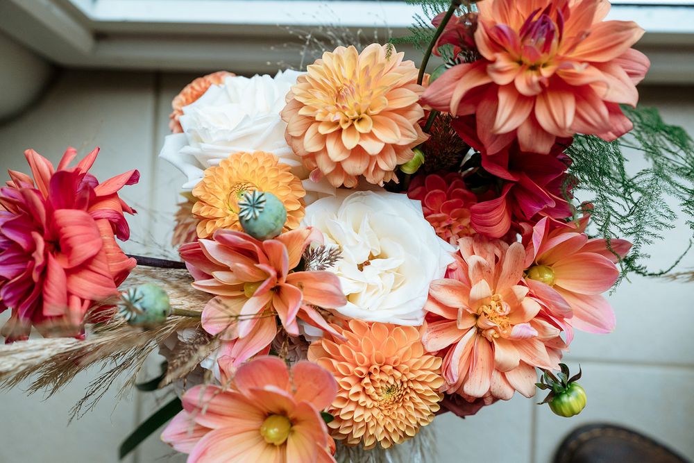 Vibrant coral and peach dahlias with white peonies arranged in a romantic floral bouquet near a window.