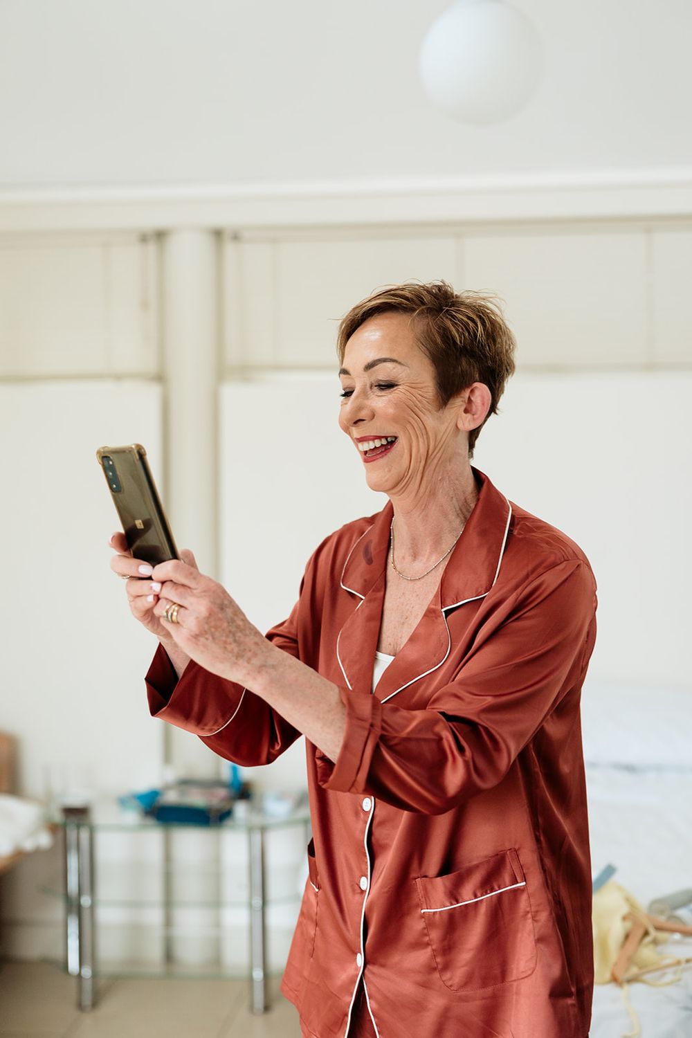 A person in a coral silk pajama set laughs while looking at their phone in a bright white room.