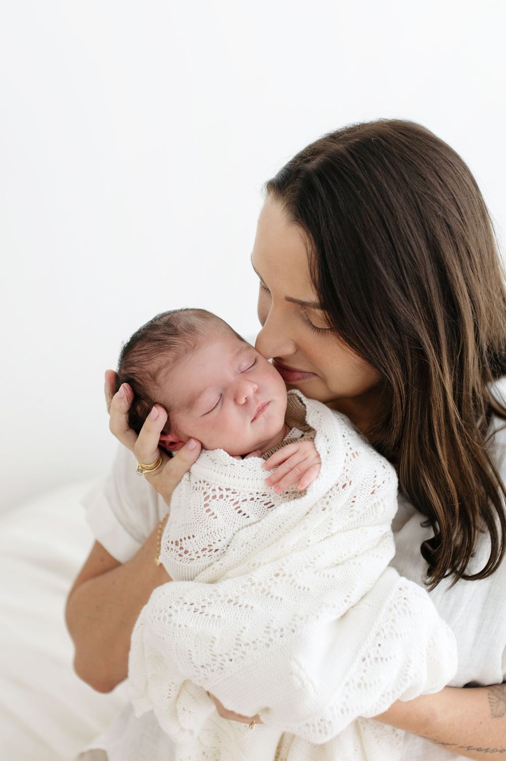A tender series of photos showing a mother holding and kissing a sleeping newborn baby in a white lace dress.