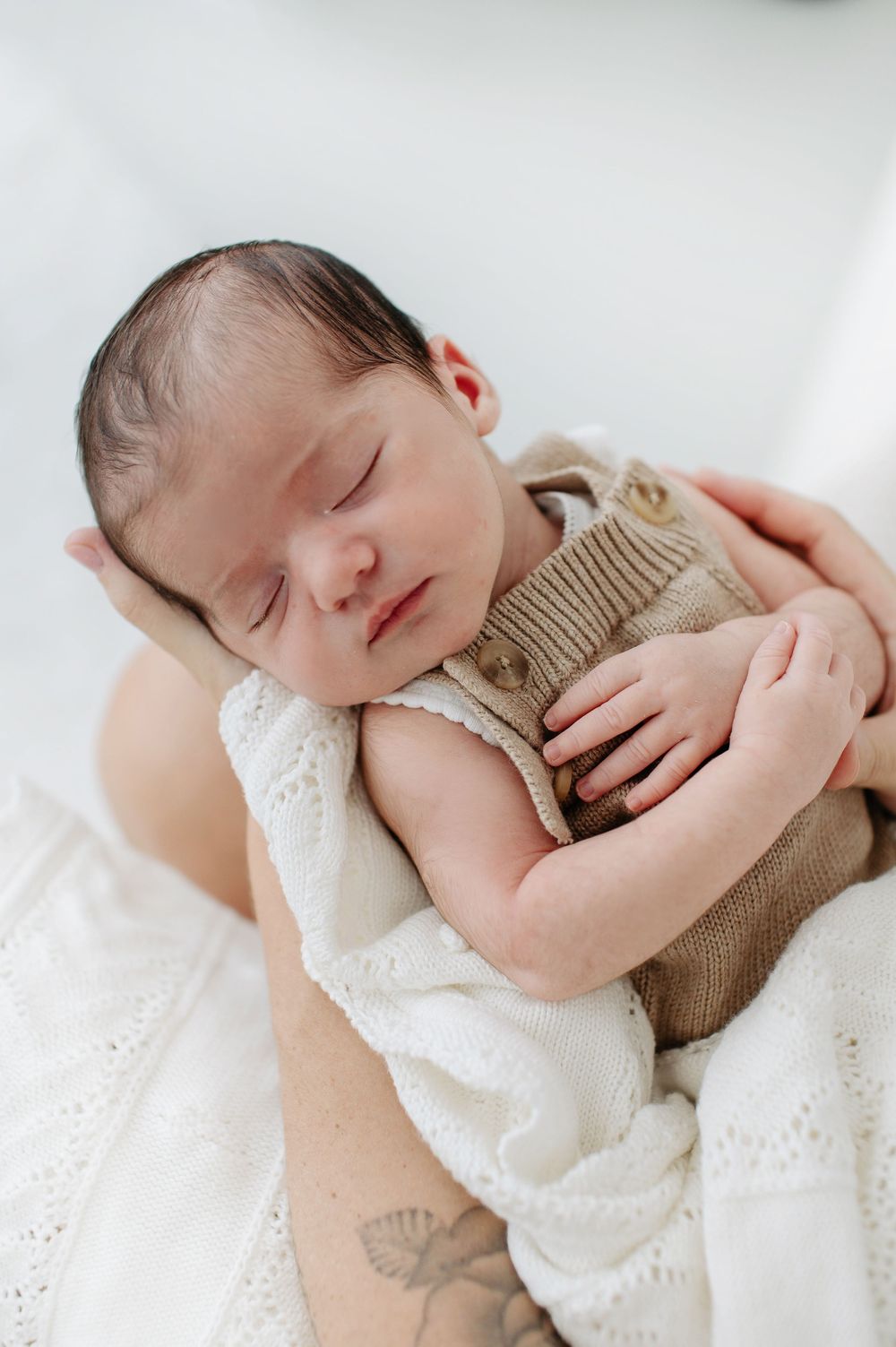 A sleeping newborn baby in a brown knit sweater rests peacefully against white lace fabric.