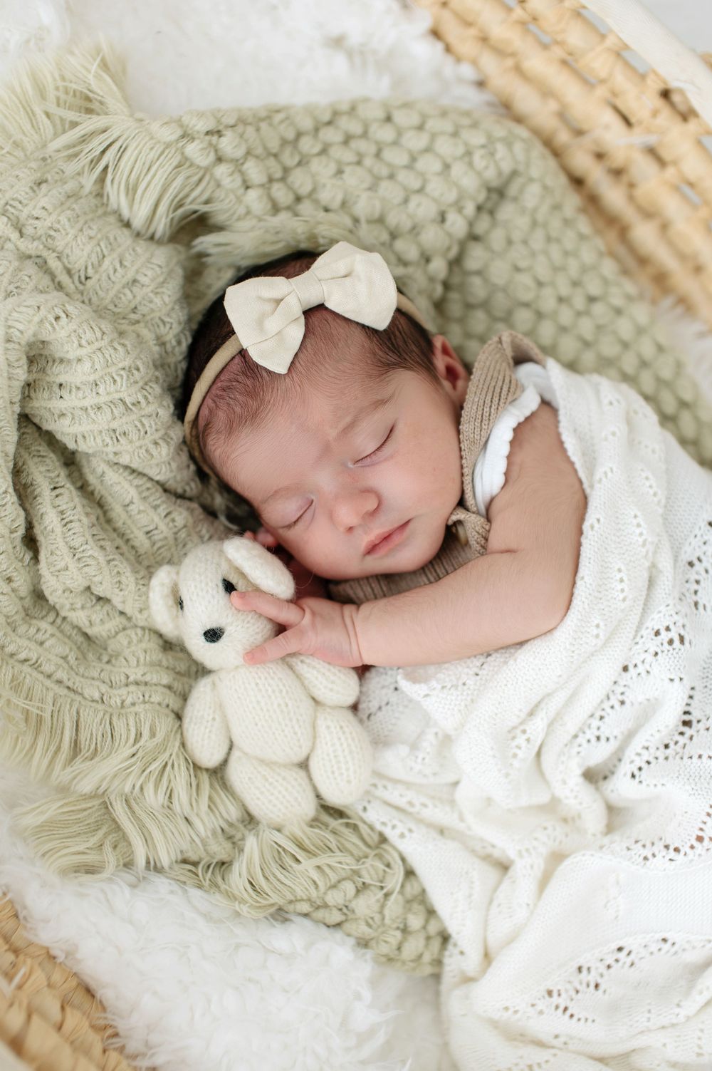 Sleeping baby in white dress with bow nestled on textured blanket with plush bunny toy.