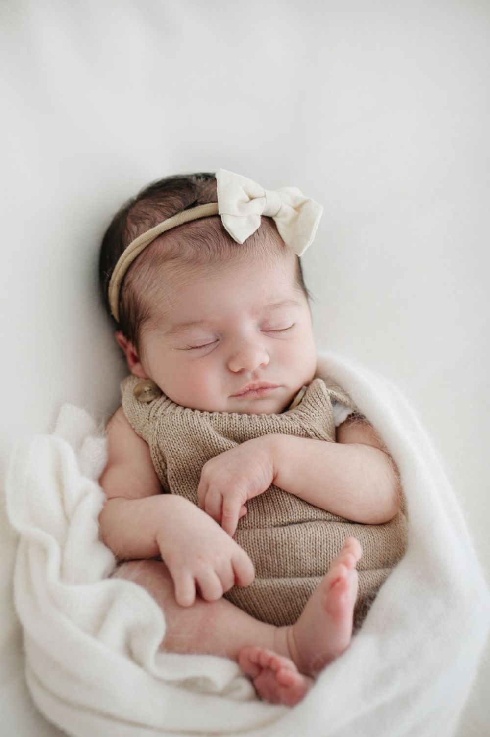 A sleeping newborn wrapped in soft white fabric wearing a knit romper during a professional photo session.