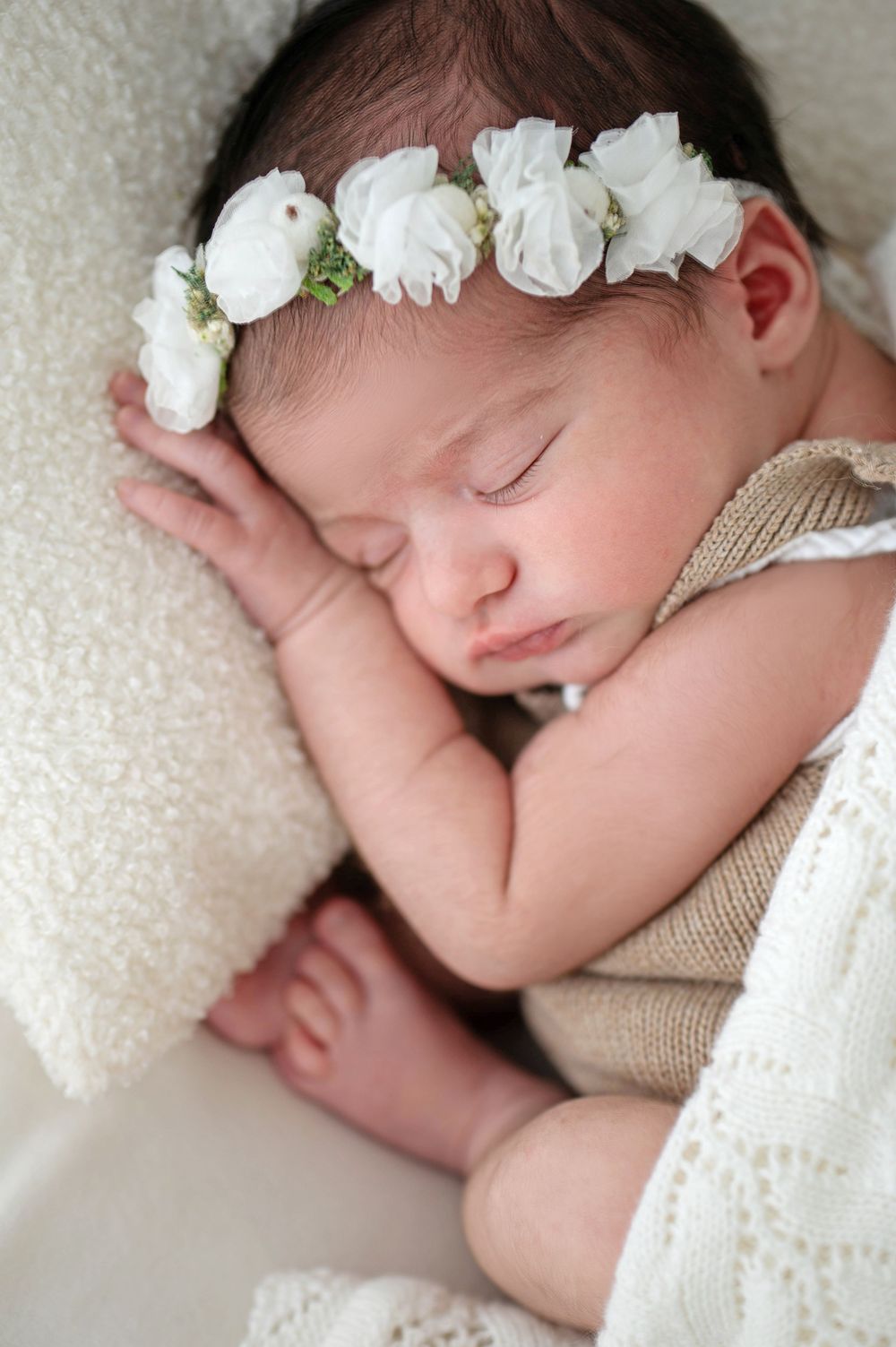 A sleeping newborn wearing a delicate white floral crown rests peacefully on cream-colored bedding.