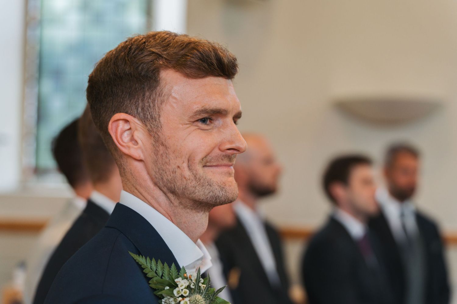 A groom in a dark suit with a boutonniere smiles warmly during his wedding ceremony.