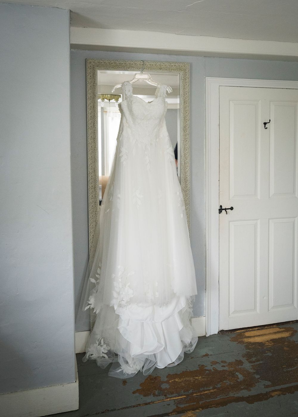 Elegant white wedding gown with lace details hanging on a mirror in a softly lit room.