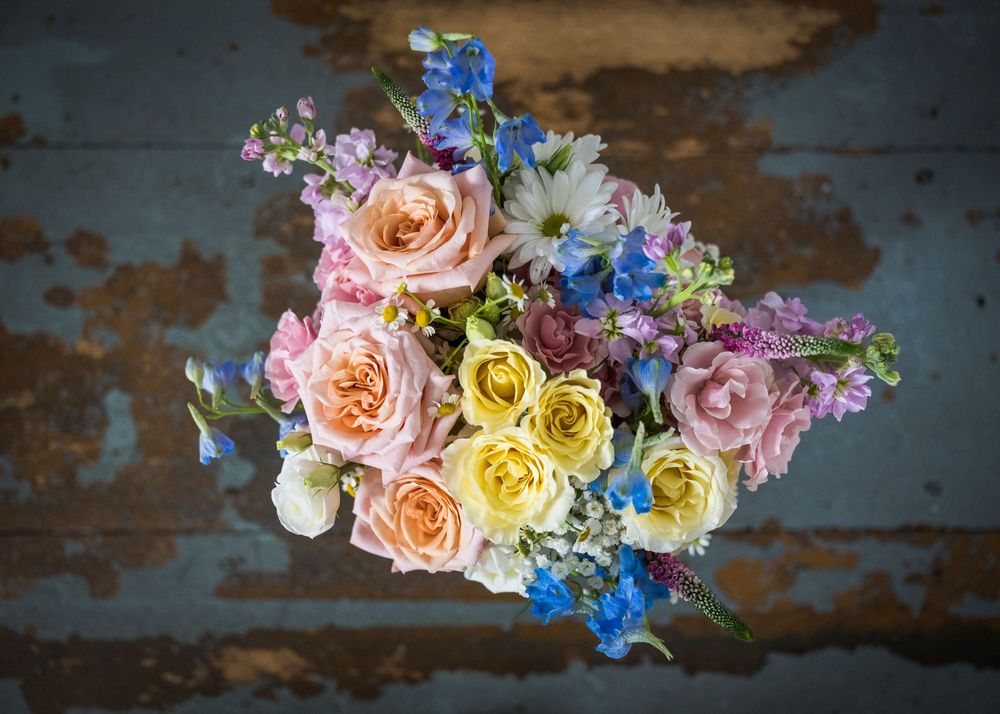 Colorful wedding bouquet with pink roses, yellow roses, blue delphiniums, and daisies on a rustic blue surface.