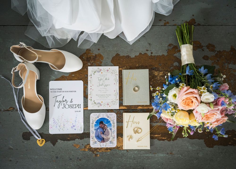 Elegant wedding flat lay featuring bridal shoes, floral bouquet, invitations, and rings on rustic surface.