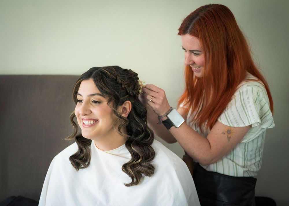 Red-haired stylist smiles while styling dark-haired bride's hair before wedding ceremony preparation.