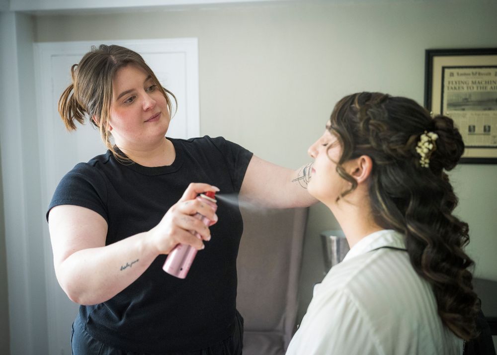 Makeup artist spraying setting spray on bride during wedding preparation in a bright room.