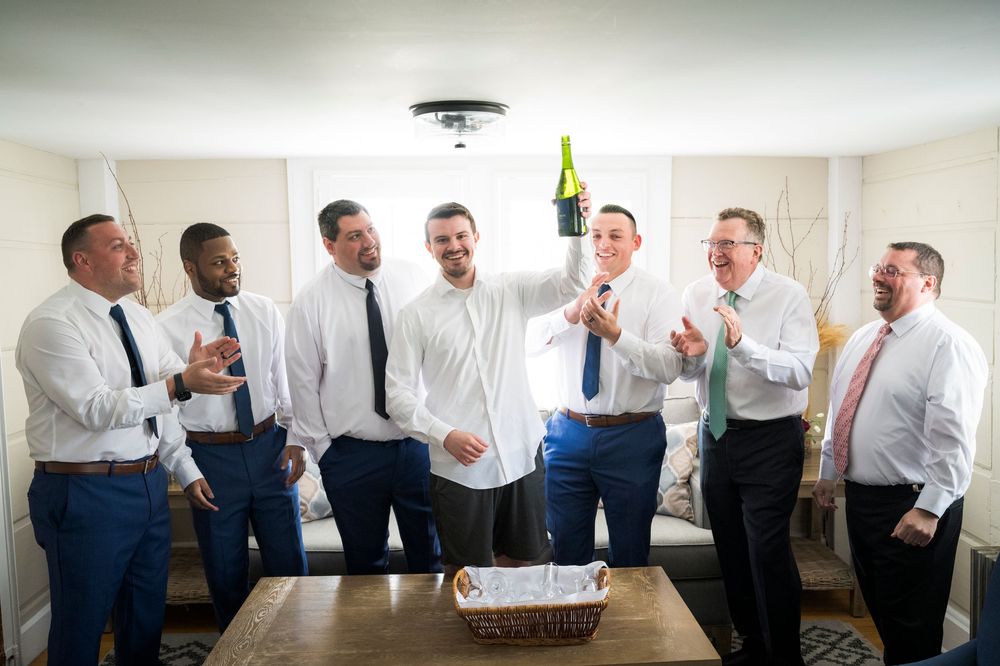 Groomsmen celebrating with a champagne bottle in a bright room before a wedding ceremony.