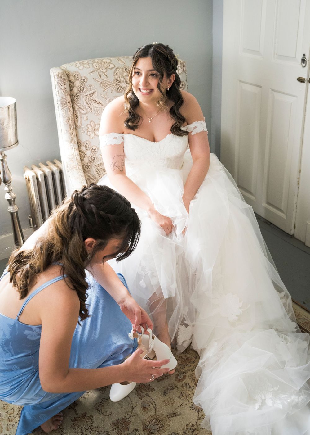 Bridesmaid in blue helps bride with shoes as she sits in floral chair in bridal suite.