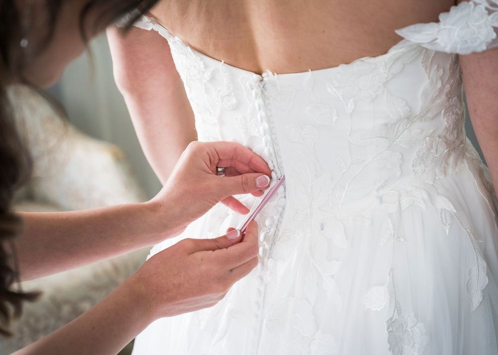 Bridesmaid fastening buttons on back of brides white lace wedding gown.