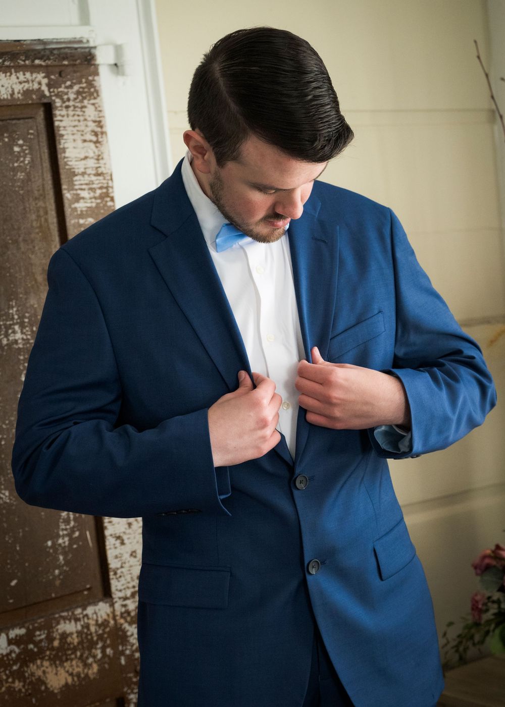 Groom in a navy blue suit and light blue bow tie adjusting his jacket before the wedding ceremony.