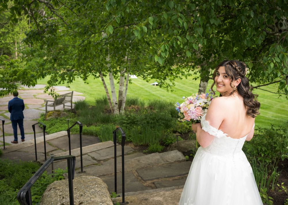 Bride in off-shoulder white gown holds pink bouquet during first look moment in lush garden setting.