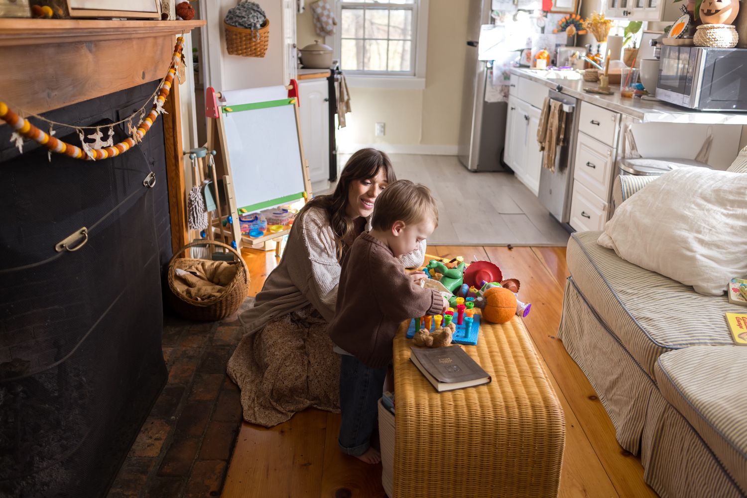 Mother wearing a cream sweater sits on wide plank wooden floor while son plays in front of fireplace for home family photos in Harvard, MA.
