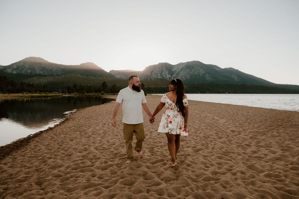 Couple walking hand in hand on a scenic beach with mountains in the background at sunset.
