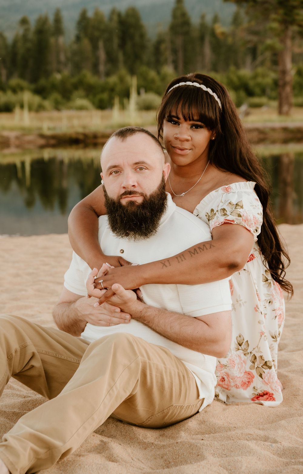 A romantic couple sits together on a sandy beach near a lake surrounded by evergreen trees in matching outfits.