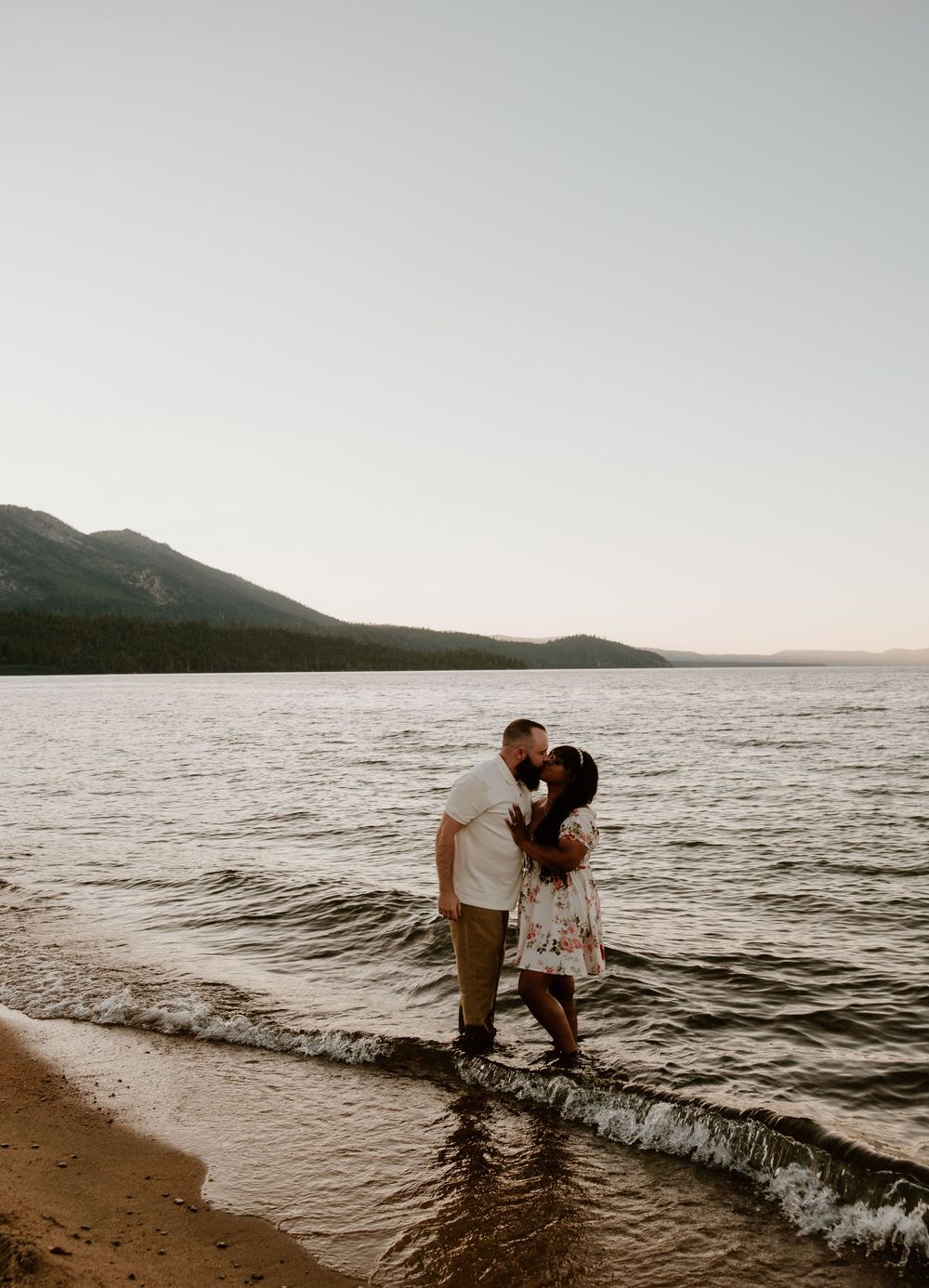 A couple shares a romantic moment on the beach at sunset with mountains in the background.