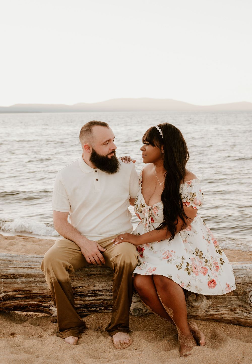 A couple sits together on driftwood at the beach during sunset, wearing casual summer attire against an ocean backdrop.