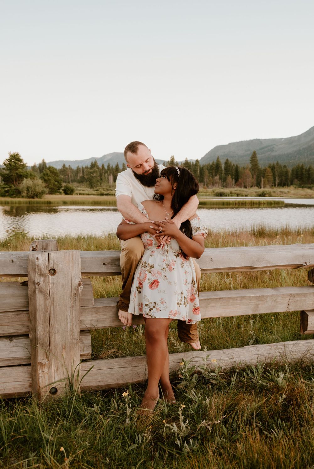 A couple embraces by a wooden fence at sunset with mountains and a lake in the background.