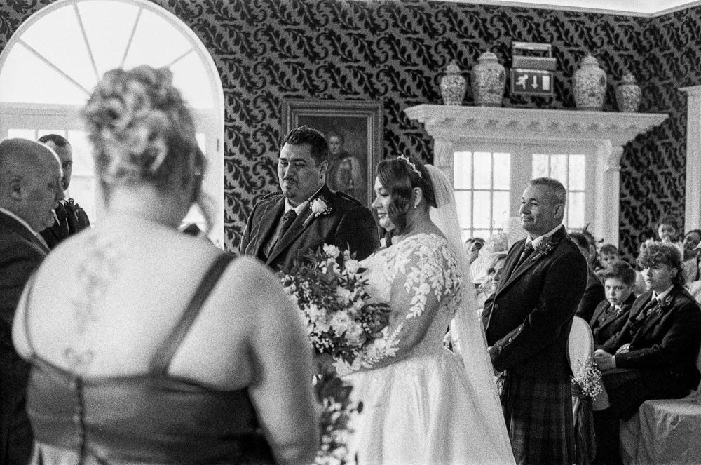 A black and white wedding ceremony photo showing a couple and guests gathered in a room with a window and decorative vases.