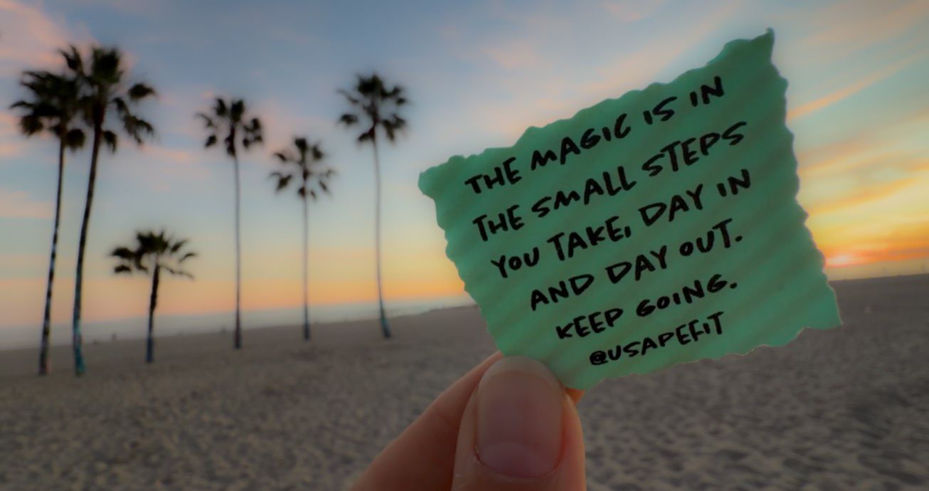 Hand holding a small green paper note against a sunset sky with palm trees silhouetted in the background.