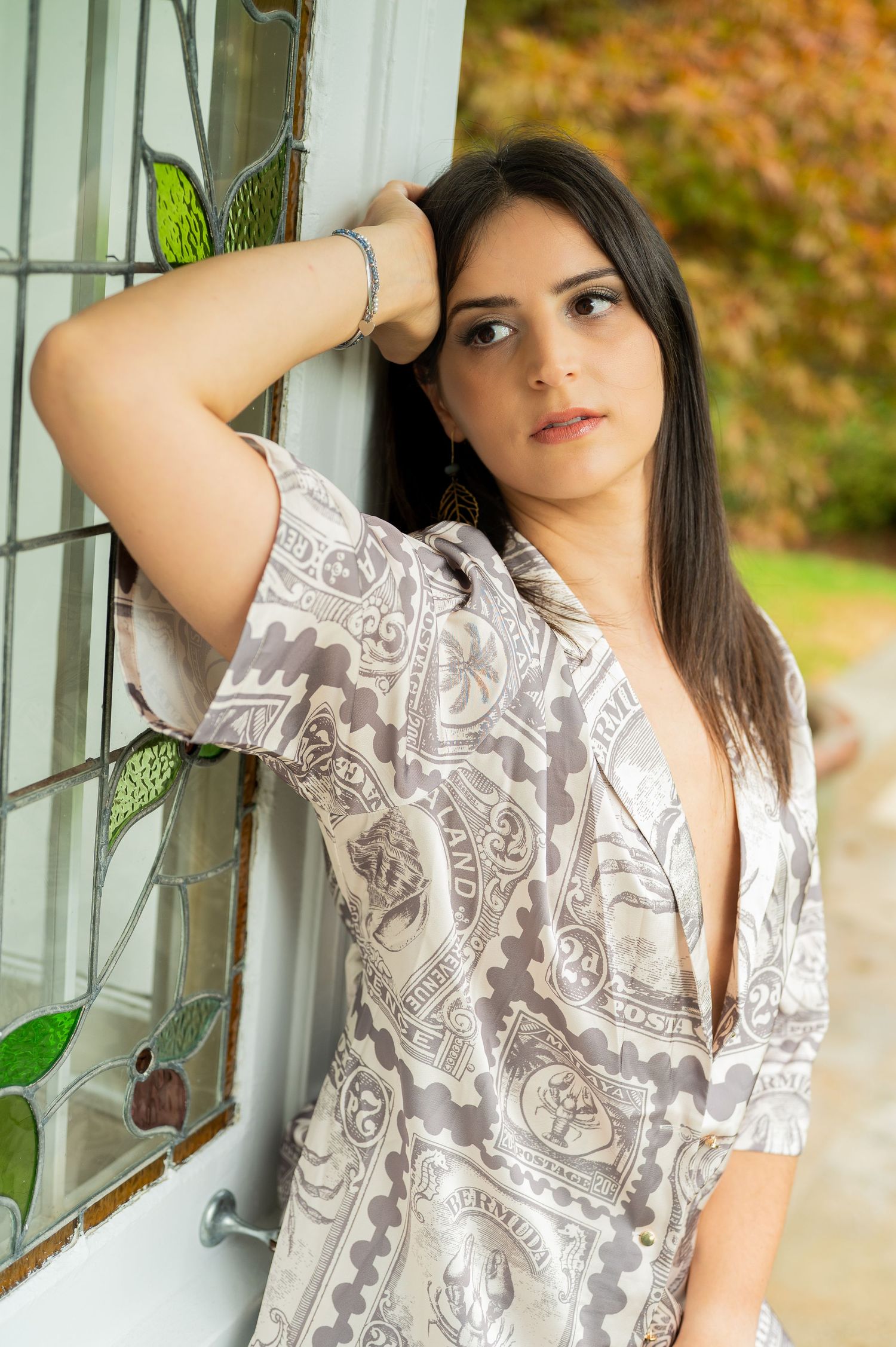 A person wearing a white and gray patterned top poses against a window with greenery in the background.