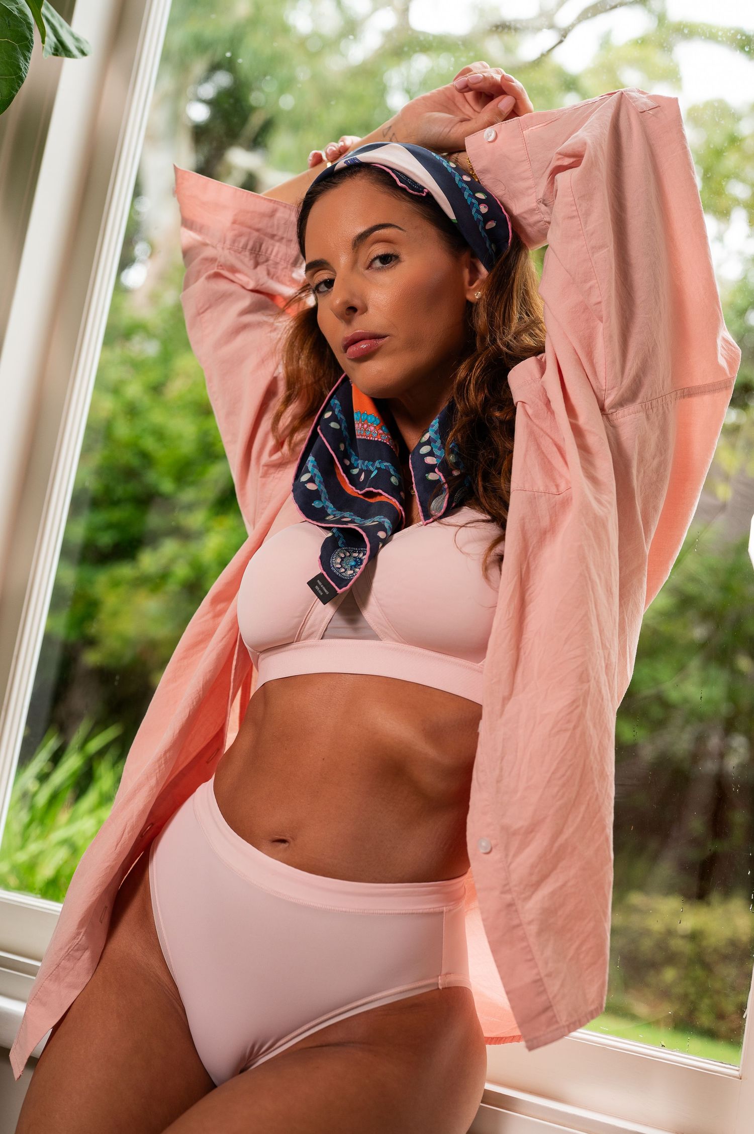 Figure in pink jacket and light undergarments poses near window with greenery outside.